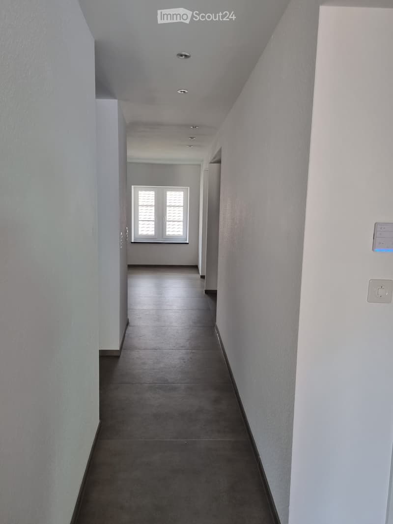 White walls, gray floor, white ceiling with recessed lighting, small window on the left wall, white door on the right wall