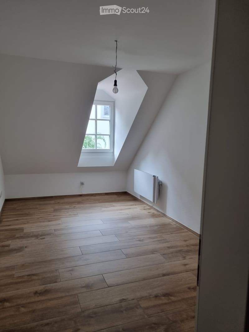 empty attic room, wooden floor, sloped ceiling, window, light fixture, wall-mounted heater, electrical outlet