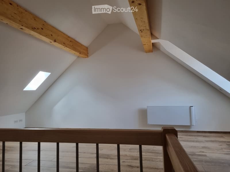 Open attic space with wooden beams, white walls, wood flooring, small skylight, and mounted radiator