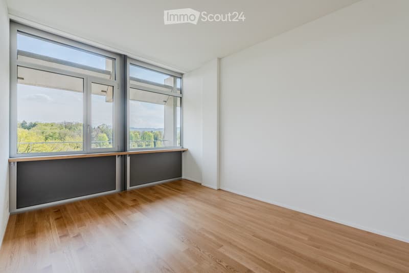 Unfurnished Room. Empty room with wooden flooring, white walls, large windows offering a view of greenery, and a dark grey radiator cover.