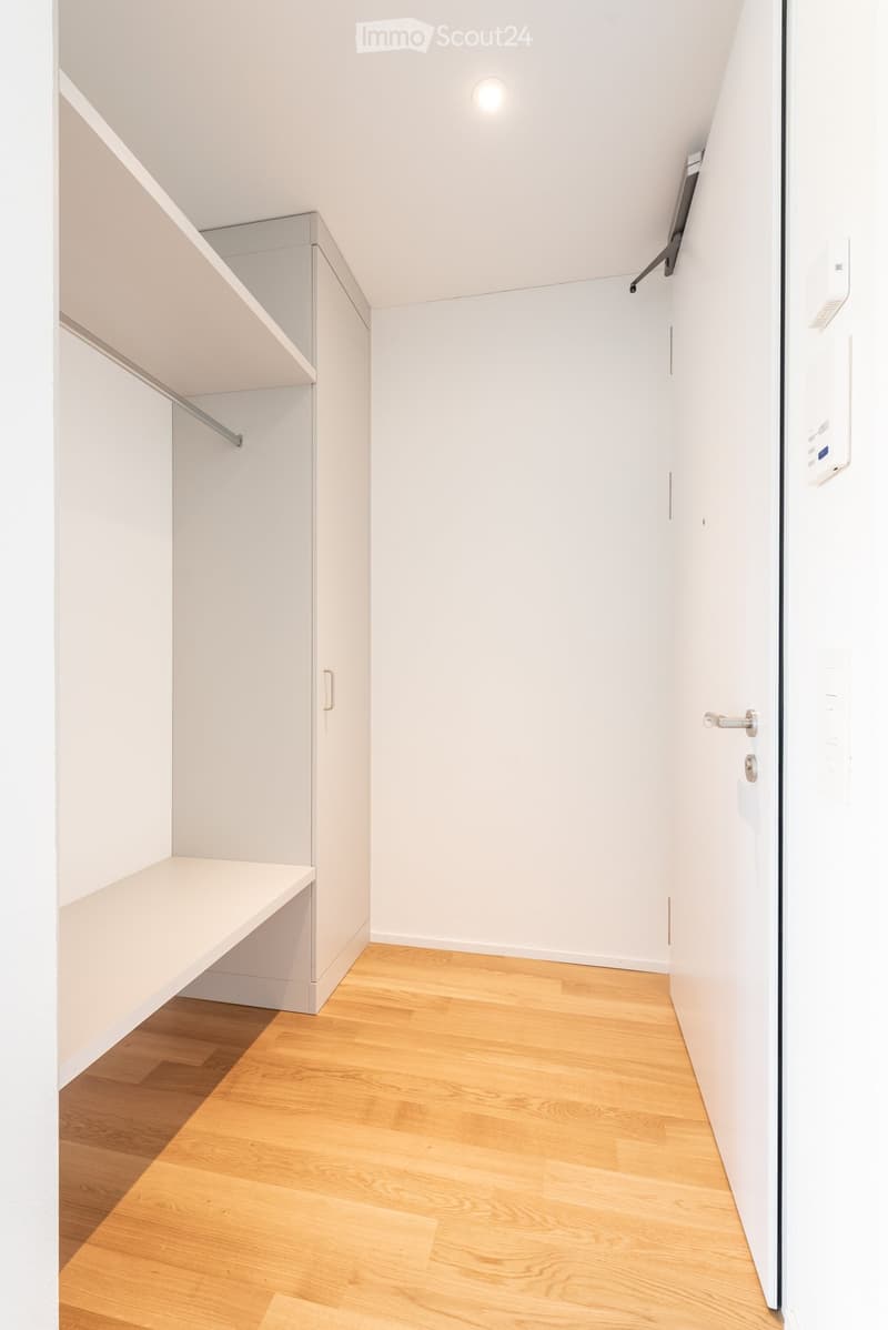 A narrow hallway with light wooden flooring, white walls, and built-in storage units on one side. The space is clean and minimalistic, with a small bench and a door at the end.