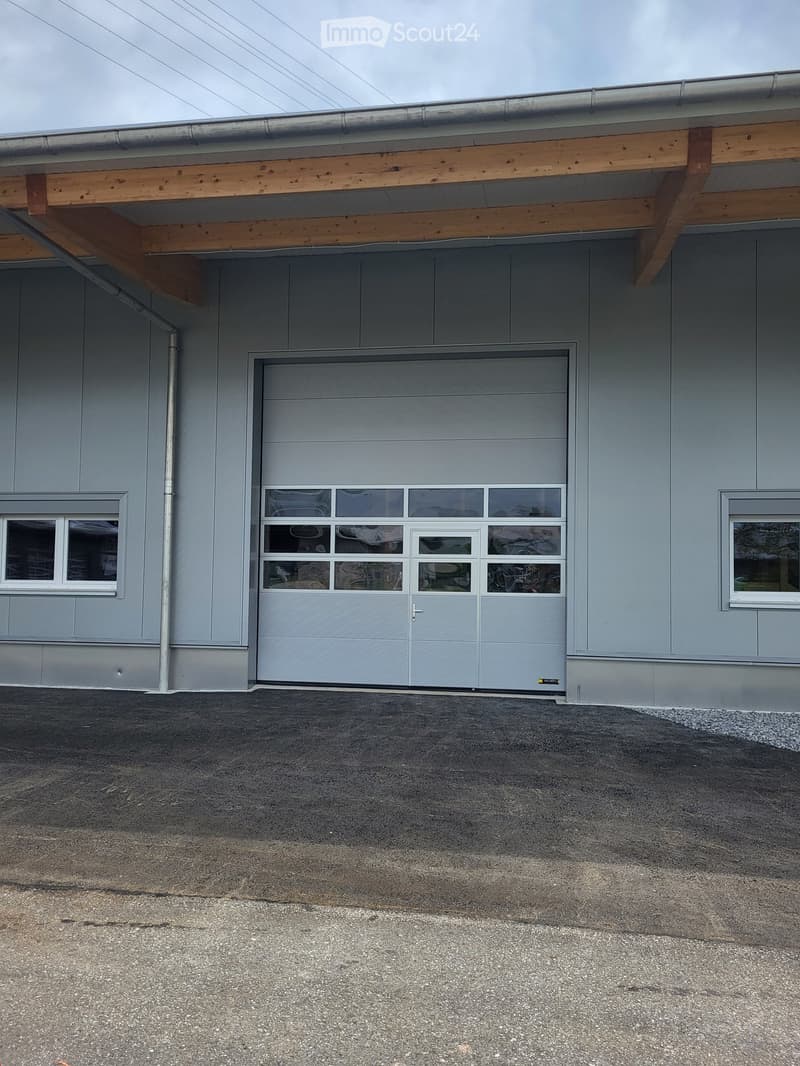 Gray building, large garage door, two small windows, metal pipe, grey gravel parking lot