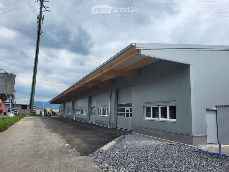 Industrial building with grey exterior, wooden beams on roof, several windows, garage doors, gravel parking area, concrete walkway