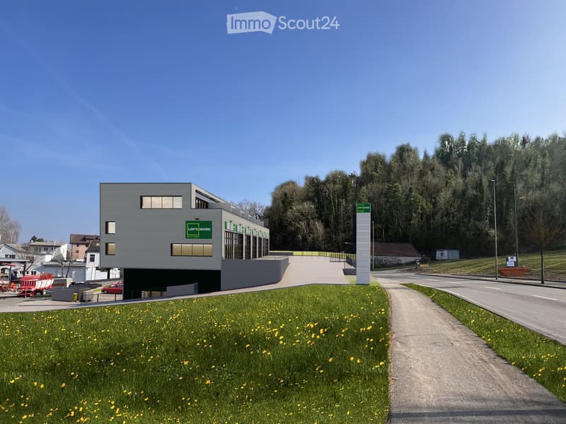 Modern two-story building with a grey exterior, windows, and green signage, situated on a sloped landscape with a pathway leading to the entrance.
