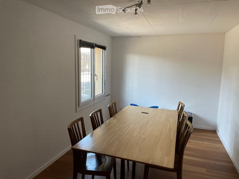 A simple interior of a room with white walls, a large wooden table, and chairs