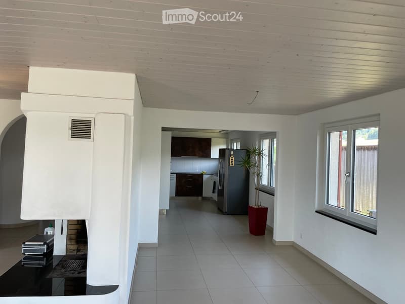 Empty room with white walls, tiled floor, a fireplace, a fridge in the kitchen, and a potted plant by the window.