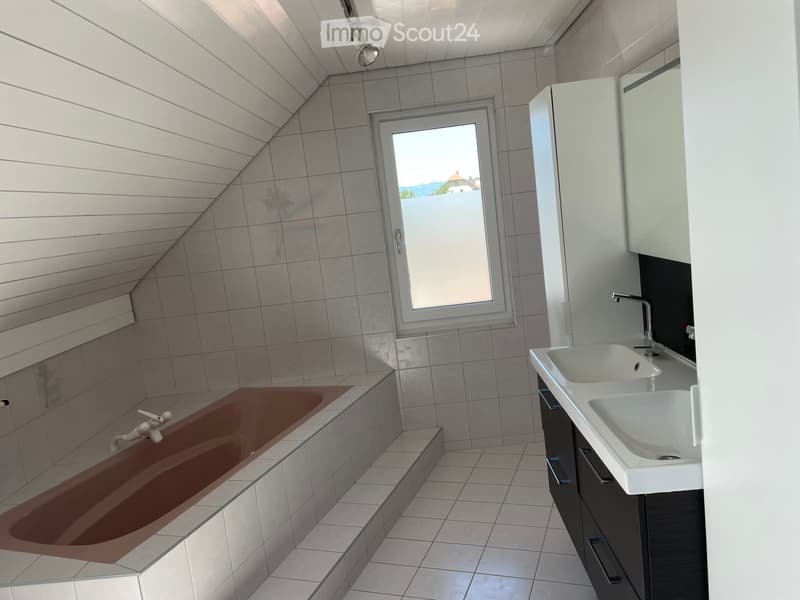 A modern bathroom featuring white walls, ceiling and tiled floor. There is a bathtub with a shower head, a window for natural light, a sink with a mirror, and modern fixtures.