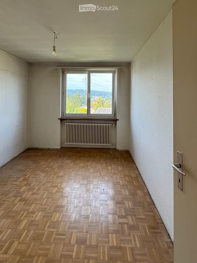 Empty room with white walls, wooden parquet floor, large window, radiator under the window, and a door.