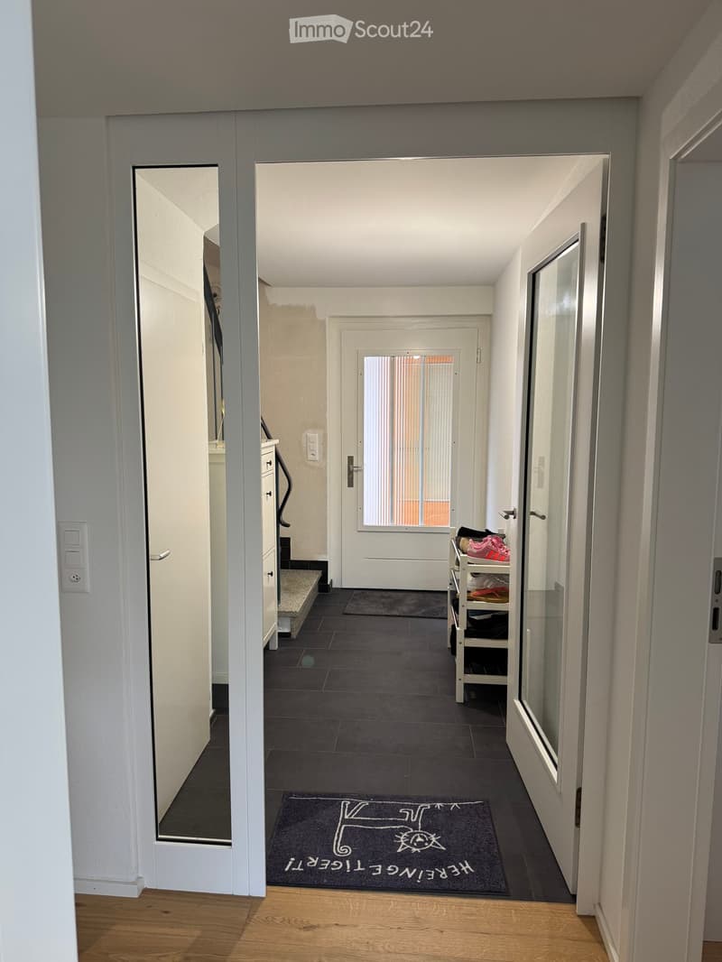 Corridor with wooden floor, white walls, glass doors, entrance mat, white door, white rack, stairs, and window