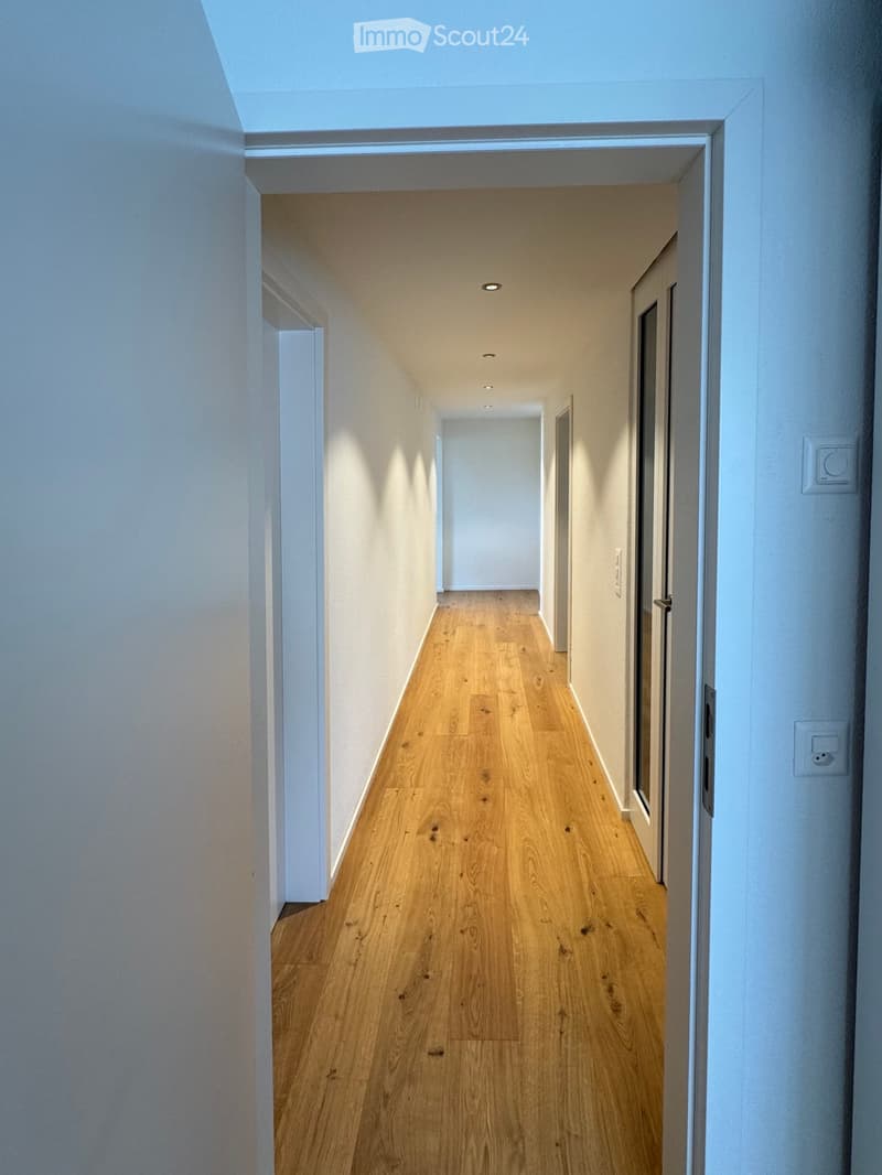 Long hallway with hardwood floors, white walls, ceiling lights, power outlets, and an open doorway at the end.