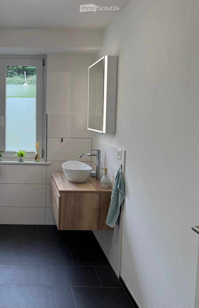 A bathroom with a modern vanity, a window overlooking a garden, white tiled walls, and a towel hanging on the wall.