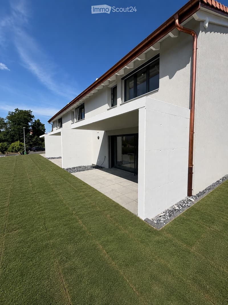 Modern two-story house with white exterior, large windows, and a covered entrance. The house features a well-maintained lawn and a paved pathway leading to the entrance.
