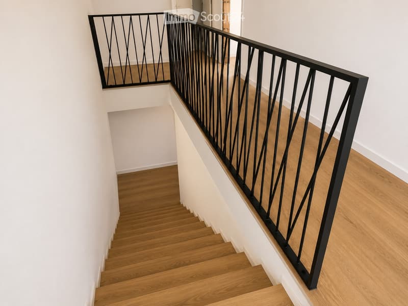 Modern staircase with wooden steps and a black metal railing featuring a geometric design. The walls are painted white, and the flooring is light wood.