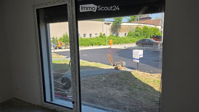 Empty room with a large window showing a view of a construction site outside, including a paved area, tools, and a car.