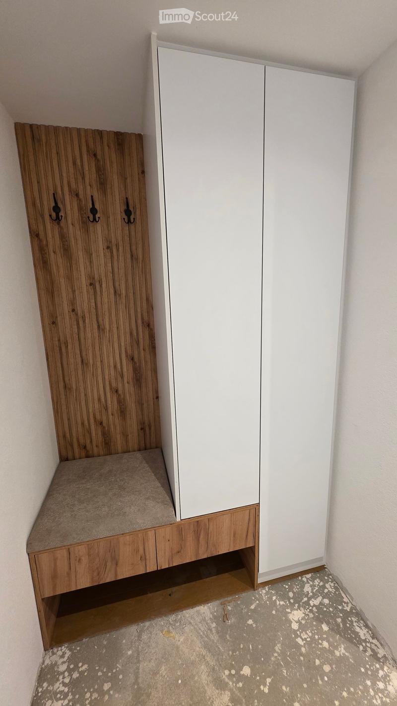 An unfurnished room with a wooden wardrobe, a built-in bench, and a white cabinet. The floor is concrete, and the walls are painted white.