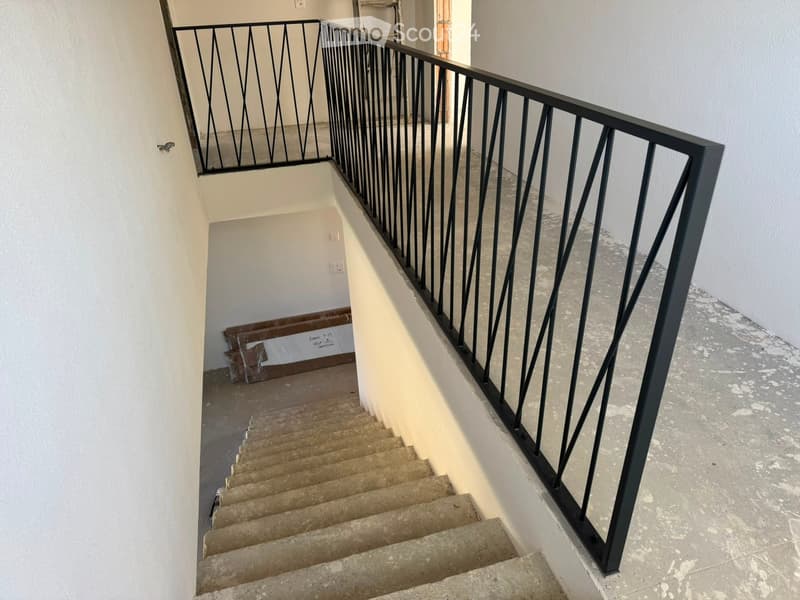 Staircase with beige steps and a black metal railing featuring a geometric pattern. The walls are white, and the floor appears to be concrete.
