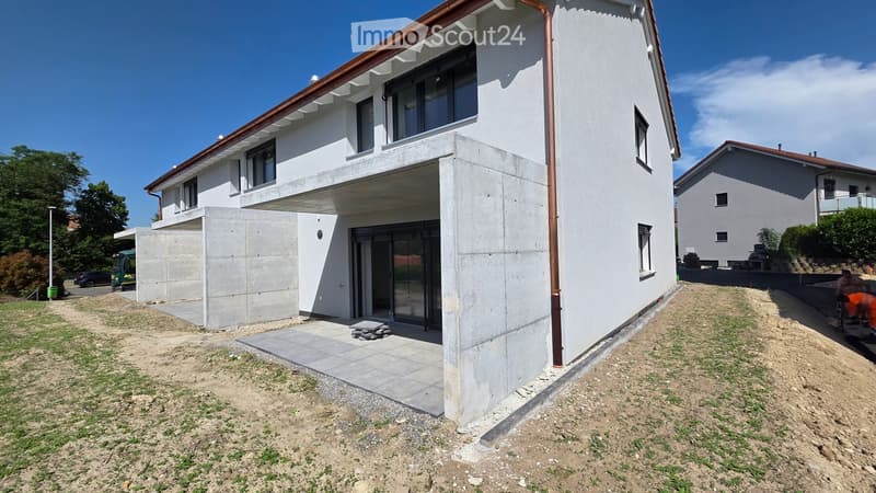 Modern two-story house with white walls, large windows, and a balcony. The house features a raised ground floor and a concrete entrance area.