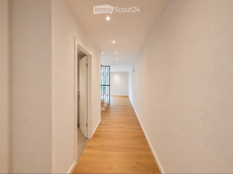 A clean, modern hallway with light wooden flooring and white walls. The corridor features recessed ceiling lighting and an open door leading to another room.