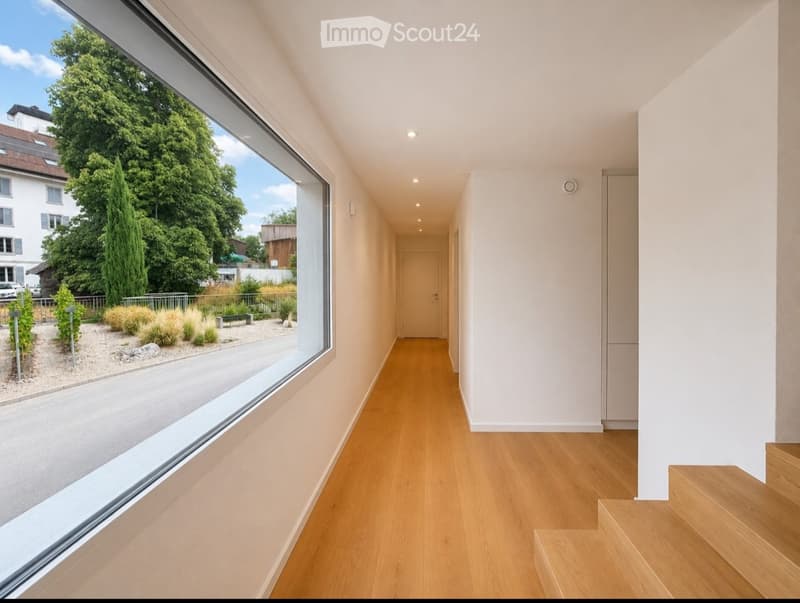 Modern hallway with light wooden flooring, white walls, recessed lighting, and a large window offering a view of the street and greenery outside.