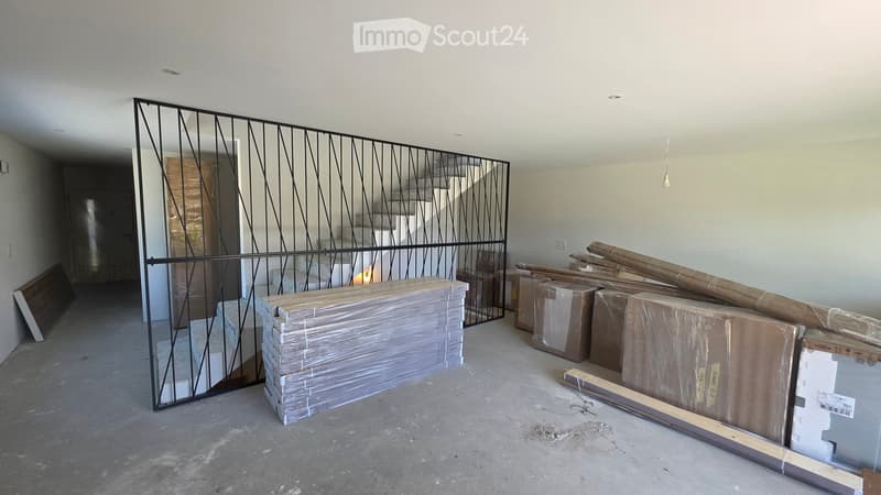 Empty room with bare walls and concrete floor, featuring a metal staircase railing and several stacked boxes and materials.