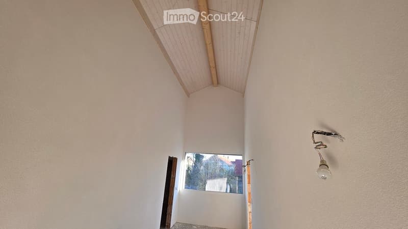 Empty room with white walls, wooden ceiling, small window, and a light fixture on the wall.