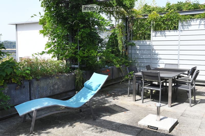 An outdoor dining area with a table and chairs, a lounge chair, and surrounded by plants and a concrete wall.