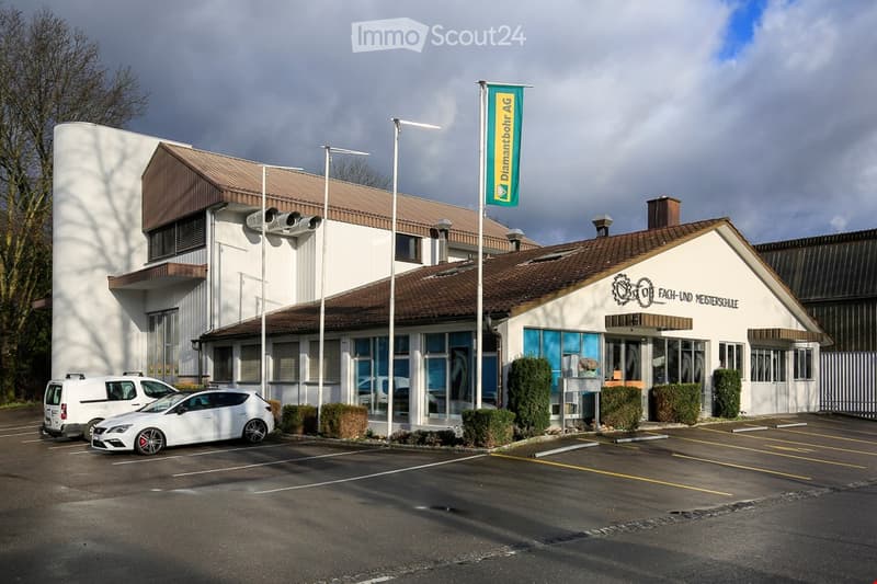 Single-storey building with a brown roof, white walls, and blue windows, multiple parking spots in front.