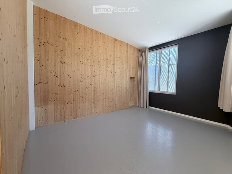 Empty room with wooden panel walls, grey carpeted floor, and a window with dark curtains.