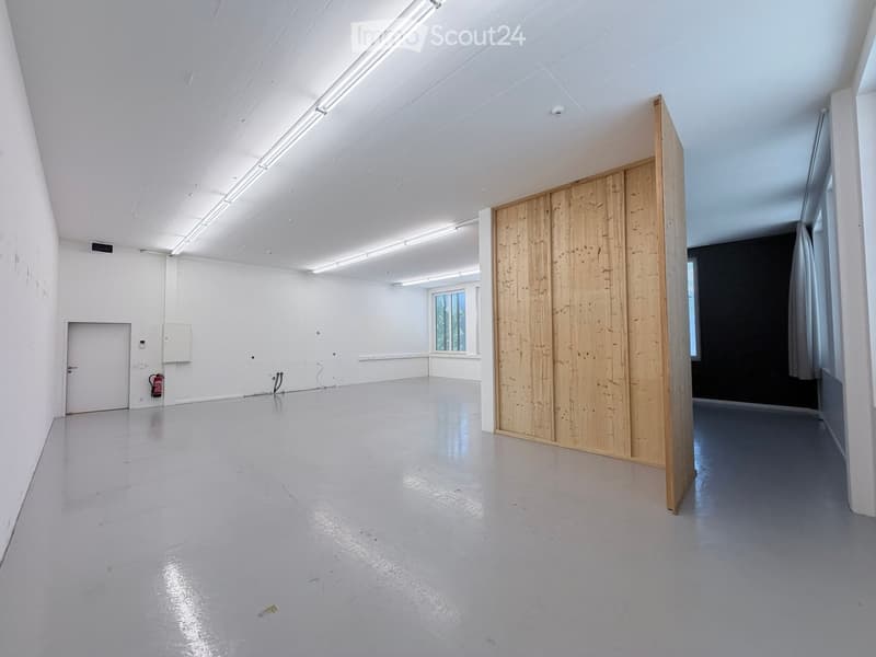 Empty room with white walls, grey floor, and a large wooden partition. Features include a door, a window, and a fire extinguisher.
