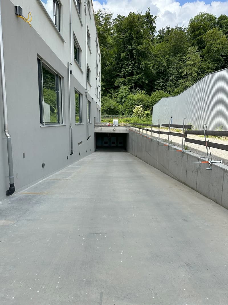 grey building exterior, concrete parking entrance, windows, pipe and cables, wooden fence, metal pole barriers, trees in the background