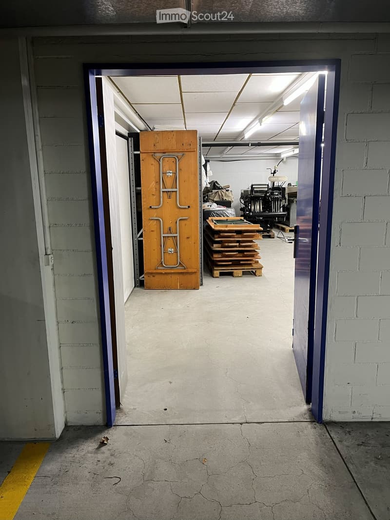 Garage entrance with open blue doors leading to an empty garage space, light fixtures on the ceiling, a stack of wooden pallets, and a large wooden door with metal bars.
