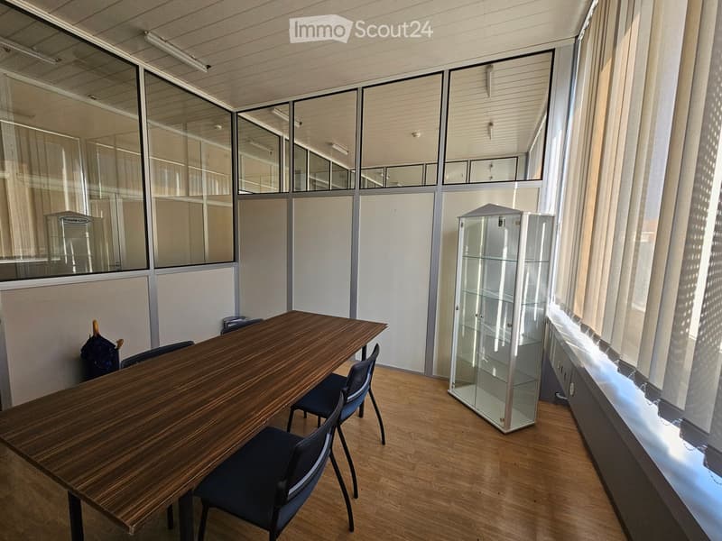 Meeting room with a long wooden table, 4 black chairs, white curtains, a glass display cabinet, ceiling mounted lighting, and a white wall with glass panels