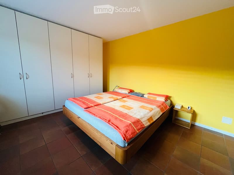 yellow wall, wooden bed with blue and orange bedding, wooden side table, white wardrobe, brown tiled floor
