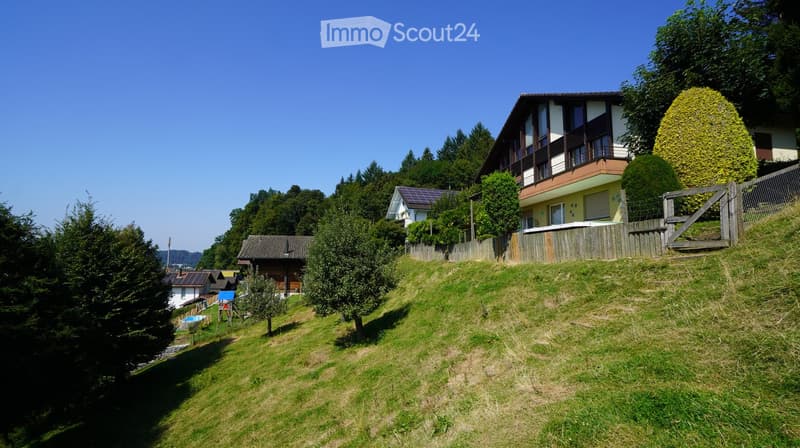 Wooden cottage on slope, solar panels, wooden fence, well-maintained garden, sunny day