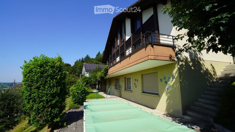 Yellow facade house, brown roof, balcony, green cover on the ground, trees and bushes on the side