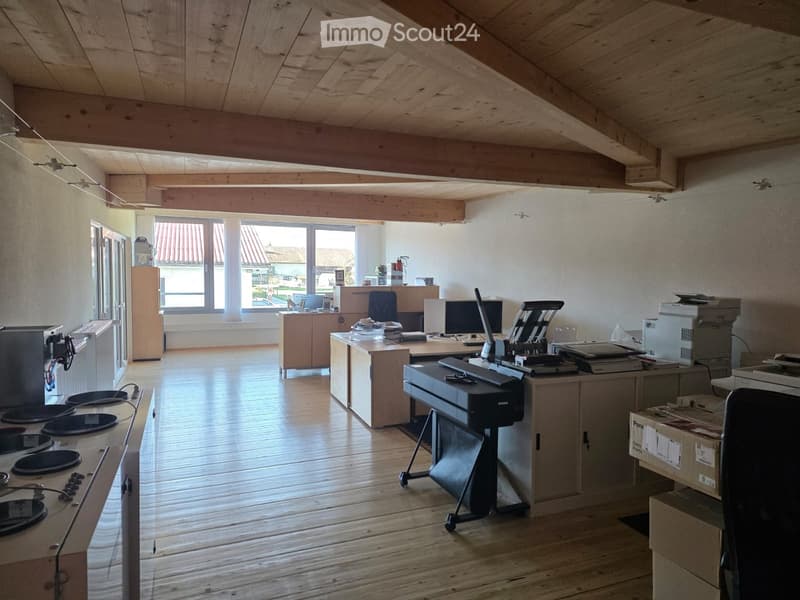 Empty office room with wooden floor, wooden beam ceiling, large windows, white walls, cabinets, computer monitors, printers, various office equipment, small office furniture, boxes