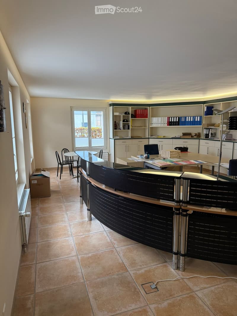 An office space featuring a reception desk, chairs, desks, shelves with organized files, a whiteboard, radiators, and a table by the window.