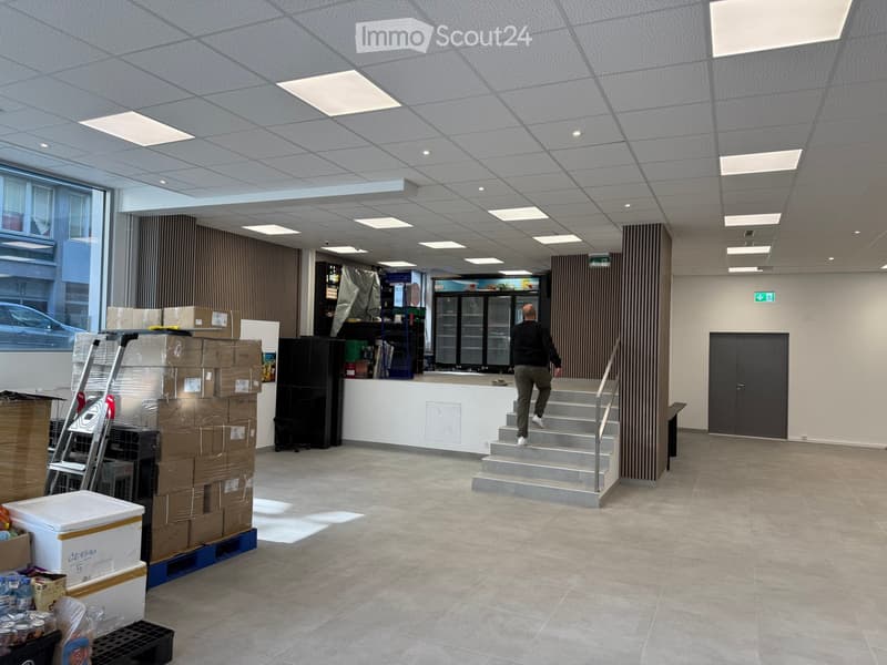 Empty room with tiled floor, white walls, ceiling with recessed lighting, a man walking up the stairs, a ladder, boxes, and a refrigerator.