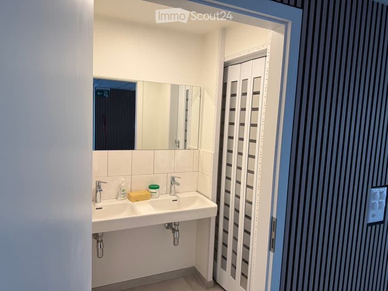 A bathroom with a tiled wall and a mirror above a sink, two sinks with faucets, and a soap dispenser on the counter.