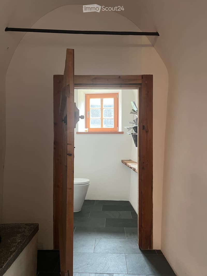 The image shows a small, white-walled bathroom with a window, toilet, and wooden door. The toilet is located near the entrance, with a wooden window frame and small decorative objects on the wall.
