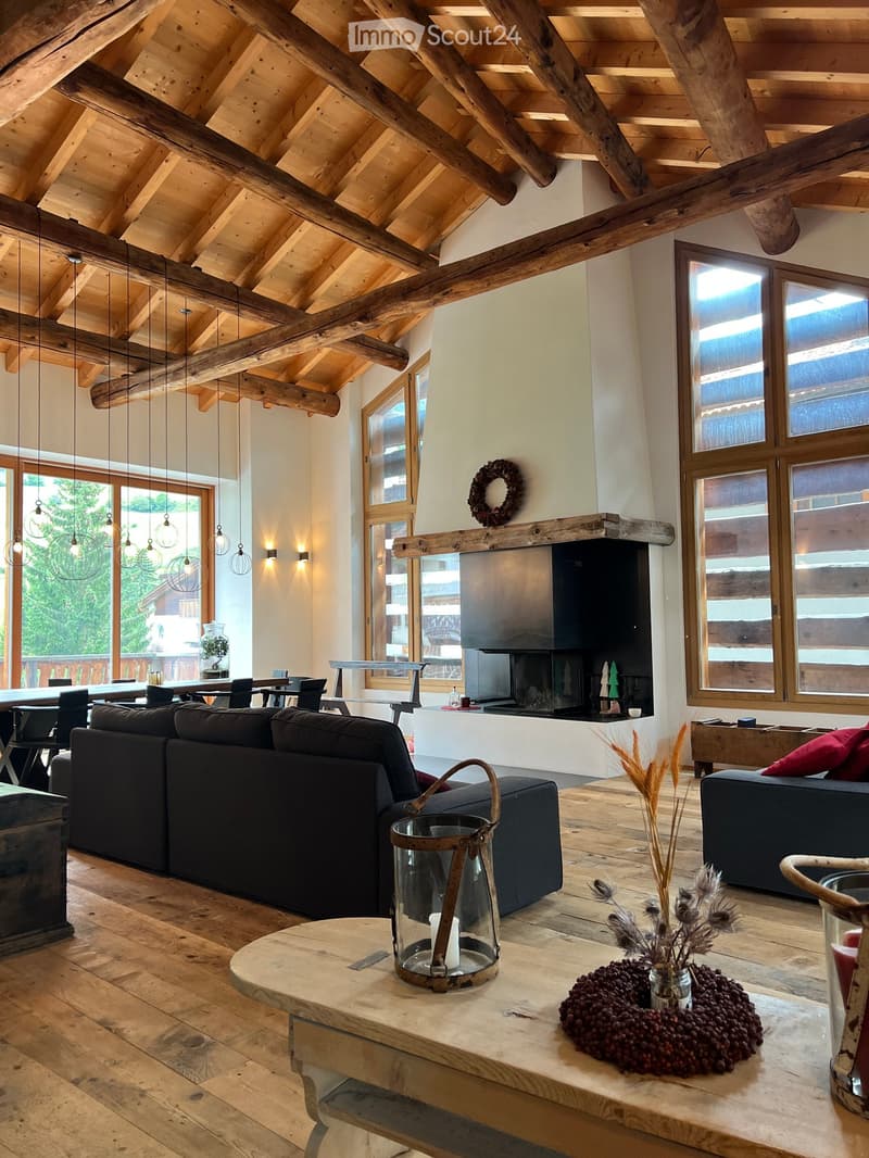 Large living room with wooden ceiling beams, black sofa, wooden floor, fireplace, large windows