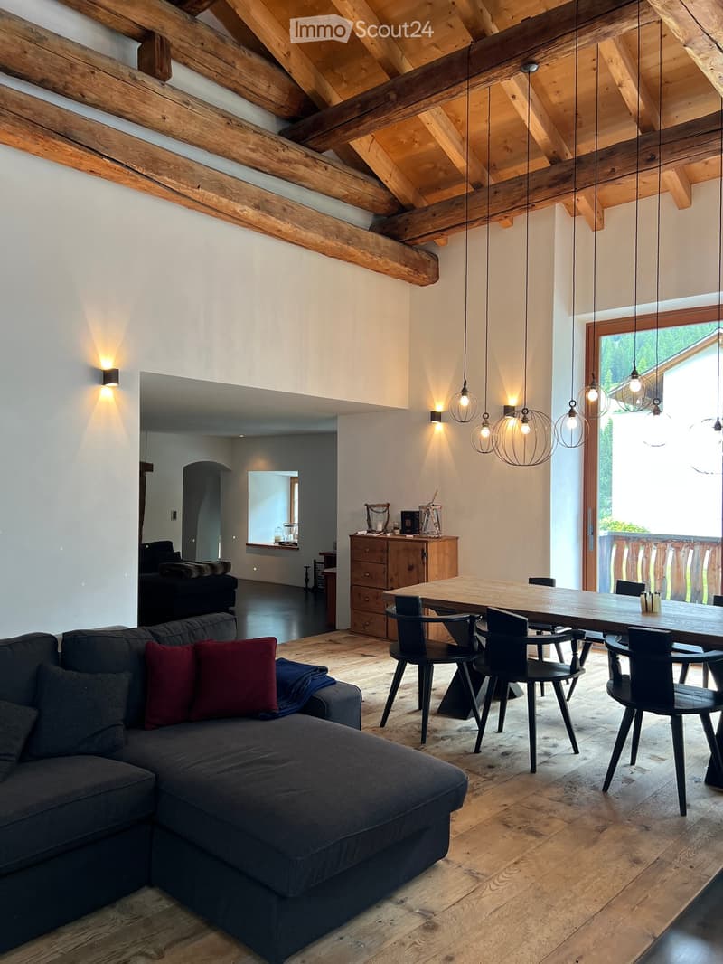 Wooden ceiling, white walls, long wooden table with chairs, couch with pillows, lamp, wooden cabinet, balcony view