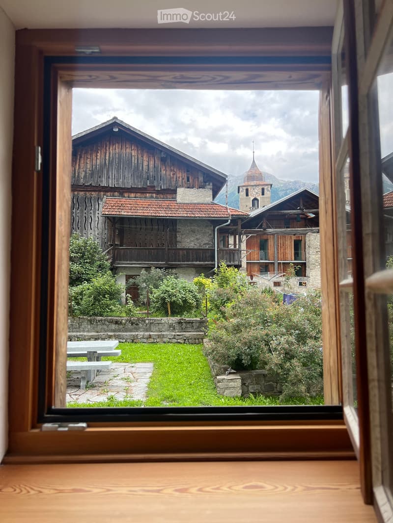 A view from a window of a traditional wooden building with a bell tower and a church in the distance, surrounded by greenery and a grassy lawn.