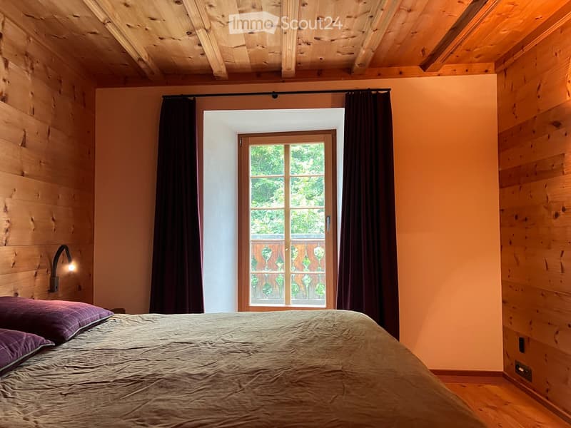 Wooden walls, bed with pillows, wall-mounted light, window with balcony, curtains