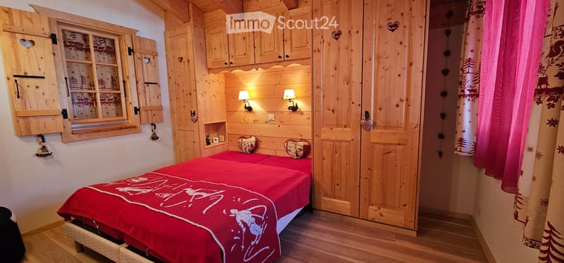 Double bed with red blanket, wooden cabin style walls, wooden floor, window with shutter, built-in wardrobes, nightstands with lamps