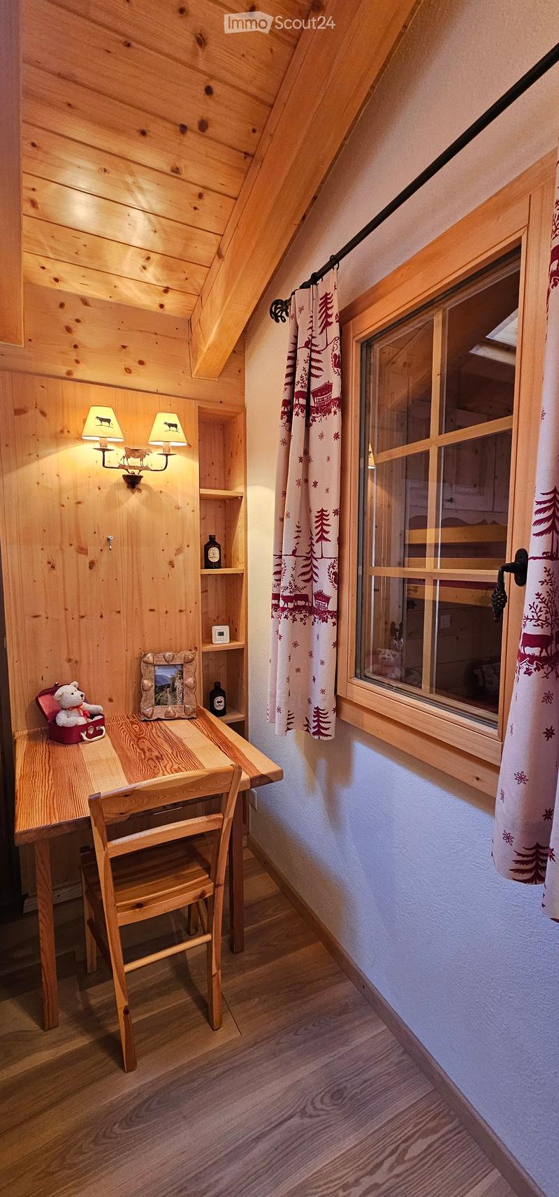 A wooden desk and chair, wooden ceiling, a wall lamp with two lamps, a small shelf with items on it, and a window with a view outside.