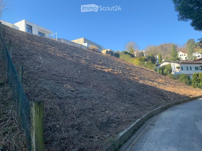 A steep hill covered with dirt and grass, adjacent to a paved road, with houses in the distance and clear blue sky above