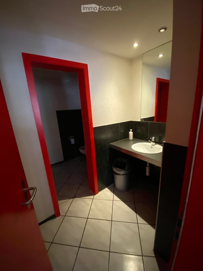 A bathroom with a sink, mirror, trash can, toilet, red trim, and white tiles.