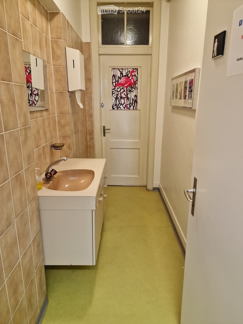 A hallway leading to a bathroom with white cabinets, a sink with a faucet, beige tiles on the wall, and a framed artwork on the wall.