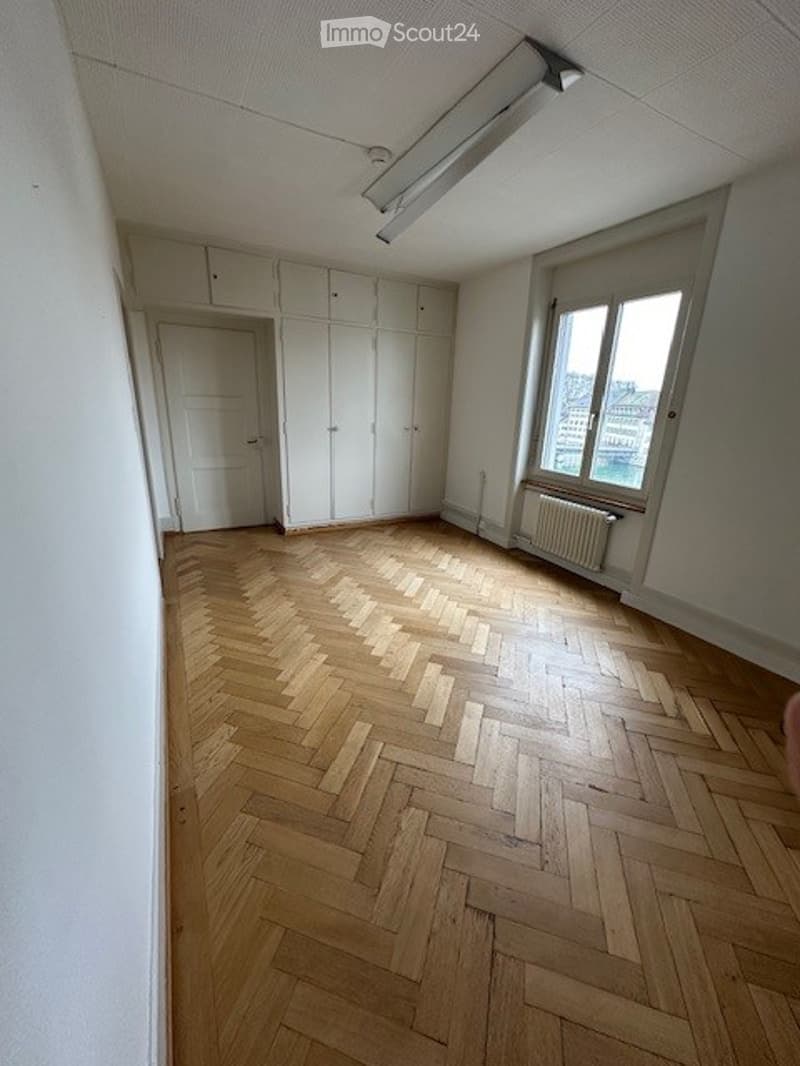 Empty room with wooden parquet floor, white walls, built-in cabinets, window with view, ceiling-mounted fluorescent lamp